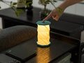 Gingko Design – Luz Twist Hexagon - Azul Laguna - Mini, lámpara de sobremesa plegable encendida, tocada por una mano, sobre una mesa de madera