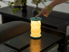 Gingko Design – Luz Twist Hexagon - Azul Laguna - Mini, lámpara de sobremesa plegable encendida, tocada por una mano, sobre una mesa de madera