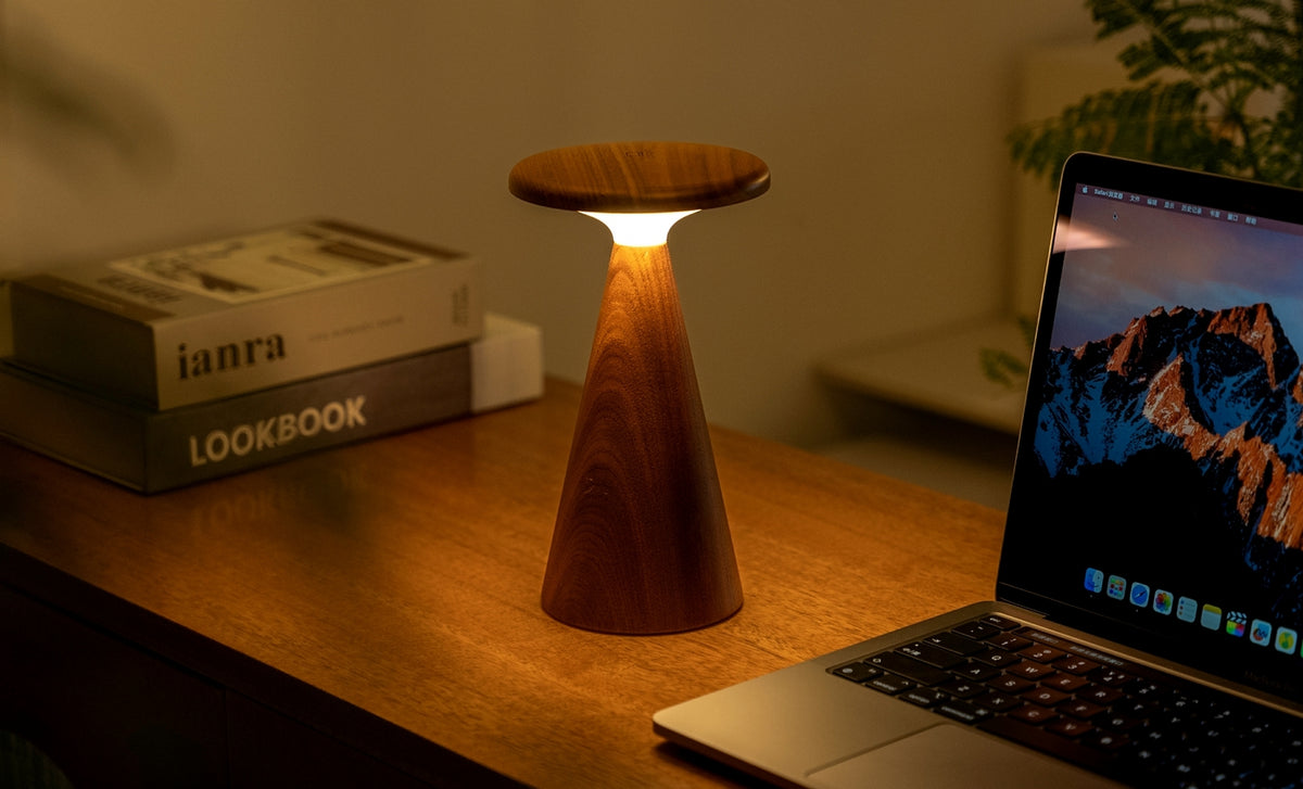 Gingko Design – Tischlampe Sylva - Walnuss - 2 Größen, eingeschaltete Holztischlampe auf einem Schreibtisch neben einem Laptop