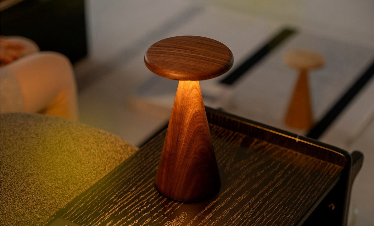 Gingko Design – Sylva Tischleuchte - Walnuss - 2 Größen, eingeschaltete Tischleuchte aus Holz auf einem Nachttisch