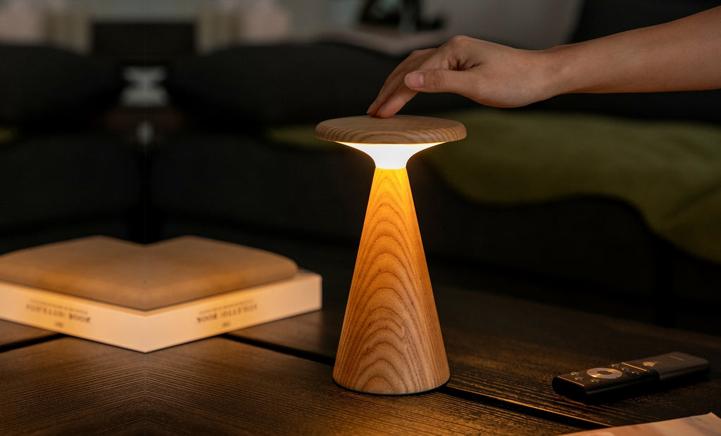 Gingko Design – Sylva Tischleuchte - Weiße Esche - 2 Größen, eingeschaltete Tischleuchte aus hellem Holz, von einer Hand berührt