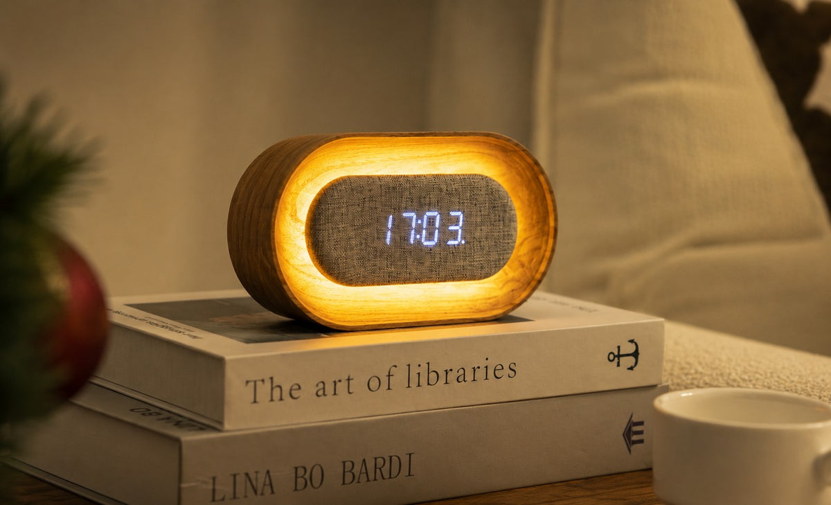 Gingko Design – Lumos Uhr - Oval Weiße Esche, Wecker aus Holz und Stoff auf Büchern platziert, warmes Licht