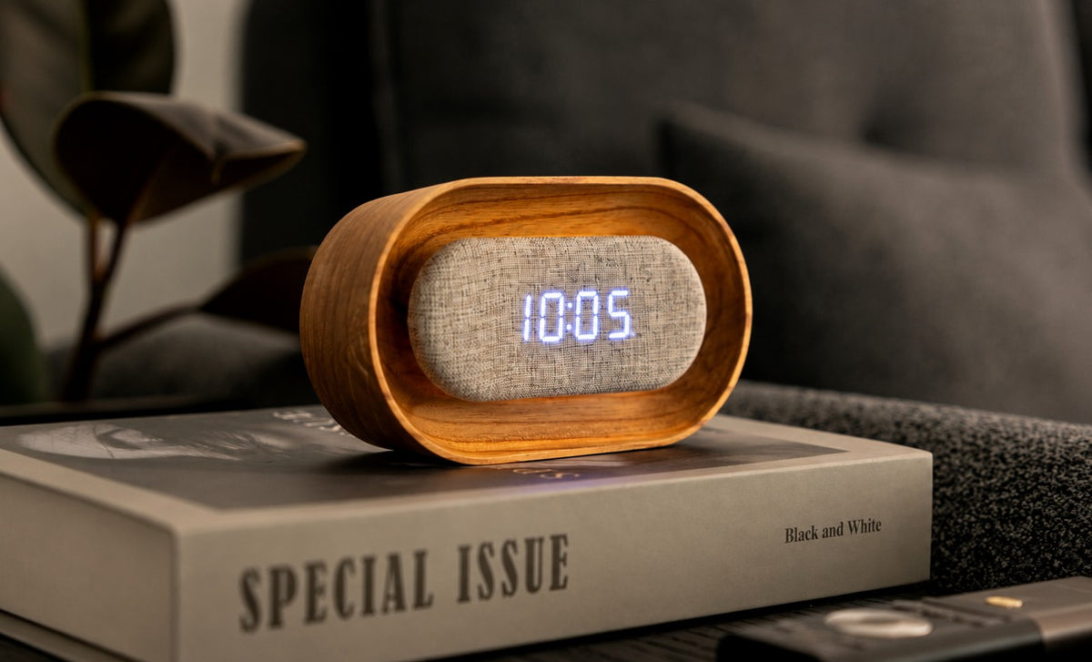 Gingko Design – Lumos Uhr - Oval Weiße Esche, Wecker aus hellem Holz und grauem Stoff auf einem Buch auf einem Nachttisch