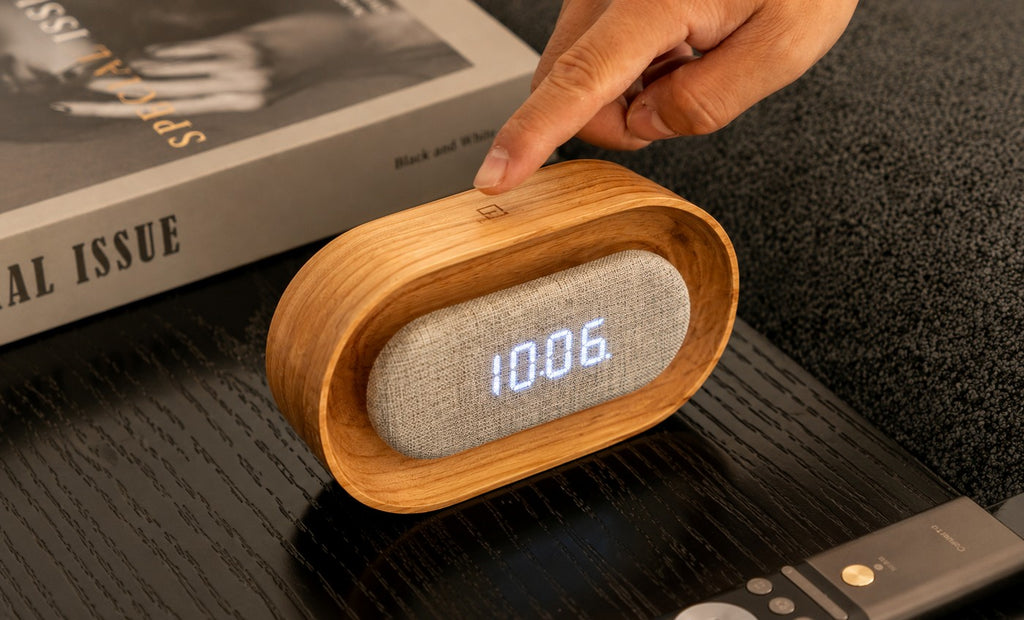 Gingko Design – Lumos Uhr - Oval Weiße Esche, digitaler Wecker aus hellem Holz und grauem Stoff auf einem Nachttisch