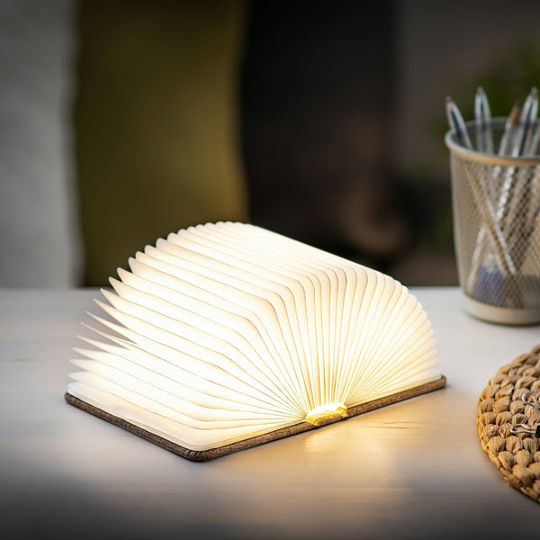 Gingko Design – Lámpara Smart Book LED - Lino Marrón Café - Mini, lámpara con forma de libro abierto, colocada sobre una mesa de madera clara
