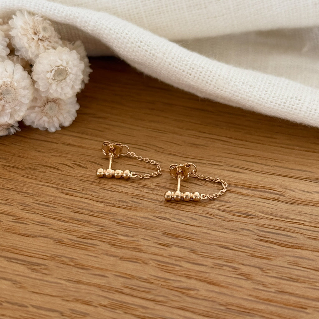 Instants Plaisirs – Boucles D'Oreilles Binta - Plaqué Or, boucles d'oreilles dorées avec chaînettes et perles sur un fond en bois