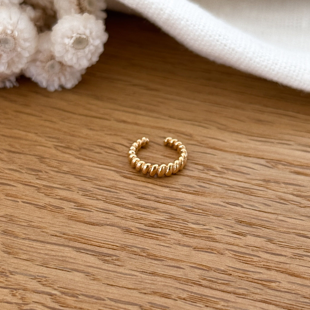 Instants Plaisirs – Bague D'Oreille Paz - Plaqué Or, bague dorée torsadée posée sur une table en bois clair