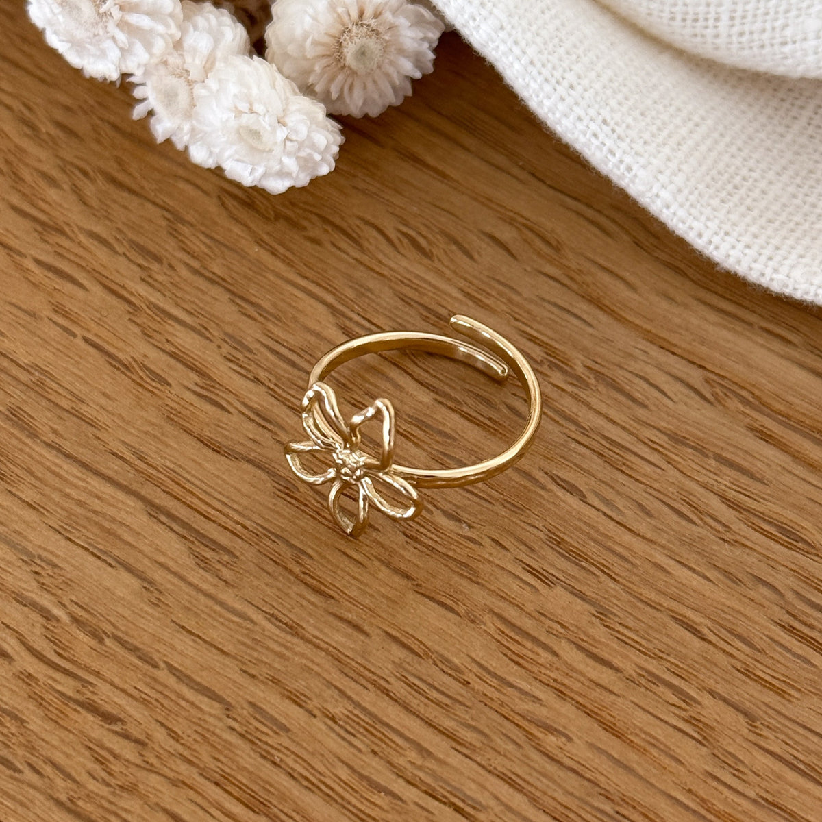 Instants Plaisirs – Bague Eline - Acier, bague dorée avec une fleur ajourée posée sur une table en bois