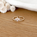 Instants Plaisirs – Bague "Adam" Plaqué Or, bague dorée avec des strass en forme de fleur, posée sur une table en bois