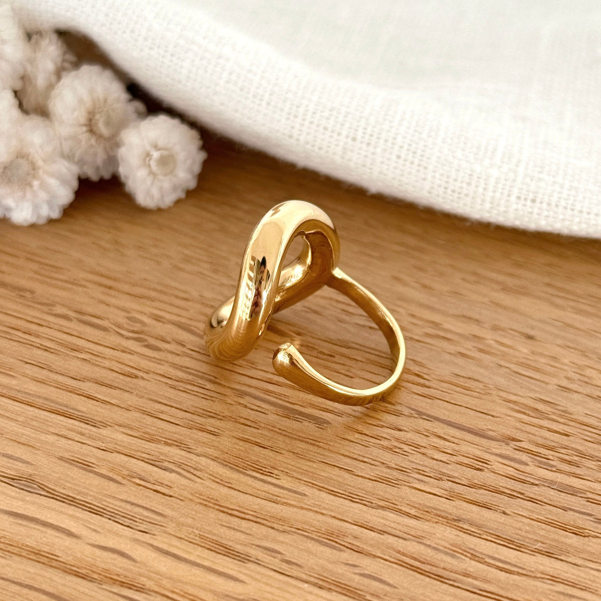 Instants Plaisirs – Bague "Orma" Acier, bague dorée en forme de boucle sur une surface en bois clair