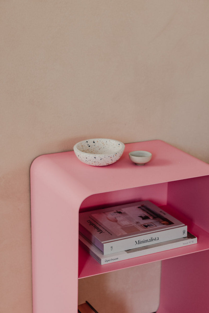 Stehender Nachttisch mit rosa Oberfläche, dekoriert mit Schalen und gestapelten Zeitschriften.