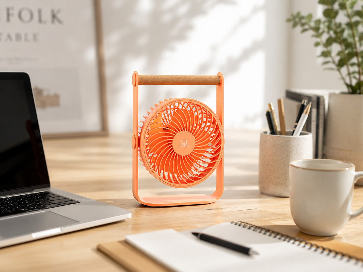 Gingko Design – Tragbarer Ventilator Lyra - Pfirsich, ein oranger Ventilator auf einem Holzschreibtisch mit einem Laptop