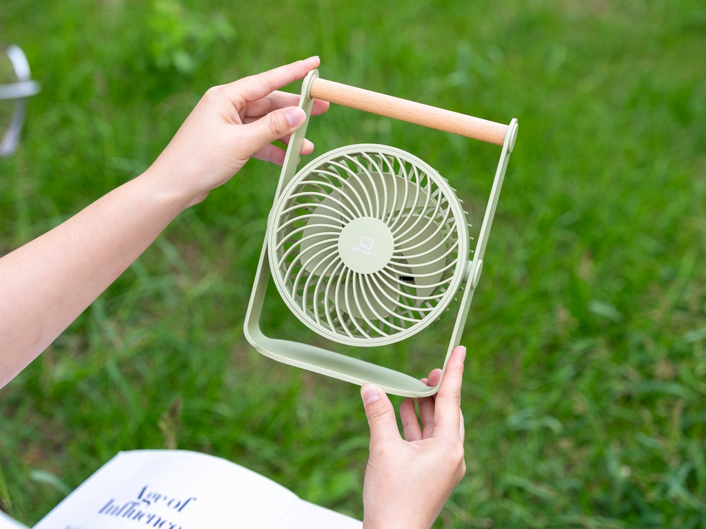 Gingko Design – Ventilador Portátil Lyra - Gris Espacial, una persona sostiene un pequeño ventilador verde claro en un jardín