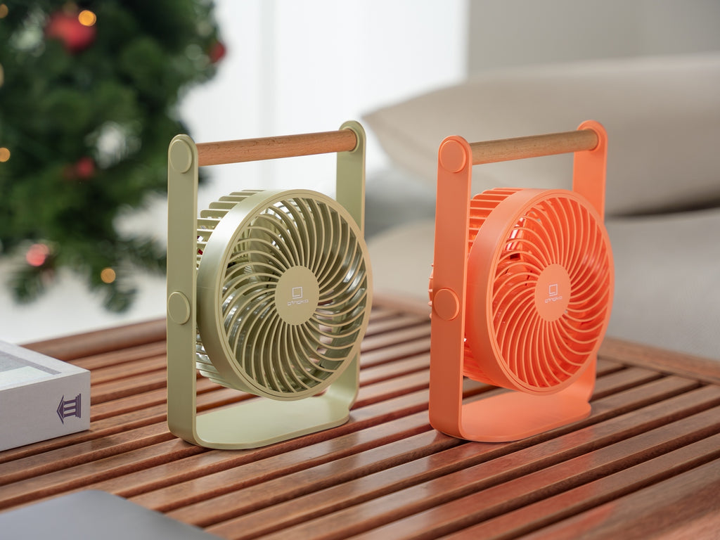 Gingko Design – Draagbare ventilator Lyra - Olijfgroen, twee draagbare ventilators, een groene en een oranje, op een houten tafel