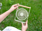 Gingko Design – Draagbare ventilator Lyra - Olijfgroen, iemand houdt een lichtgroene kleine ventilator vast in een tuin