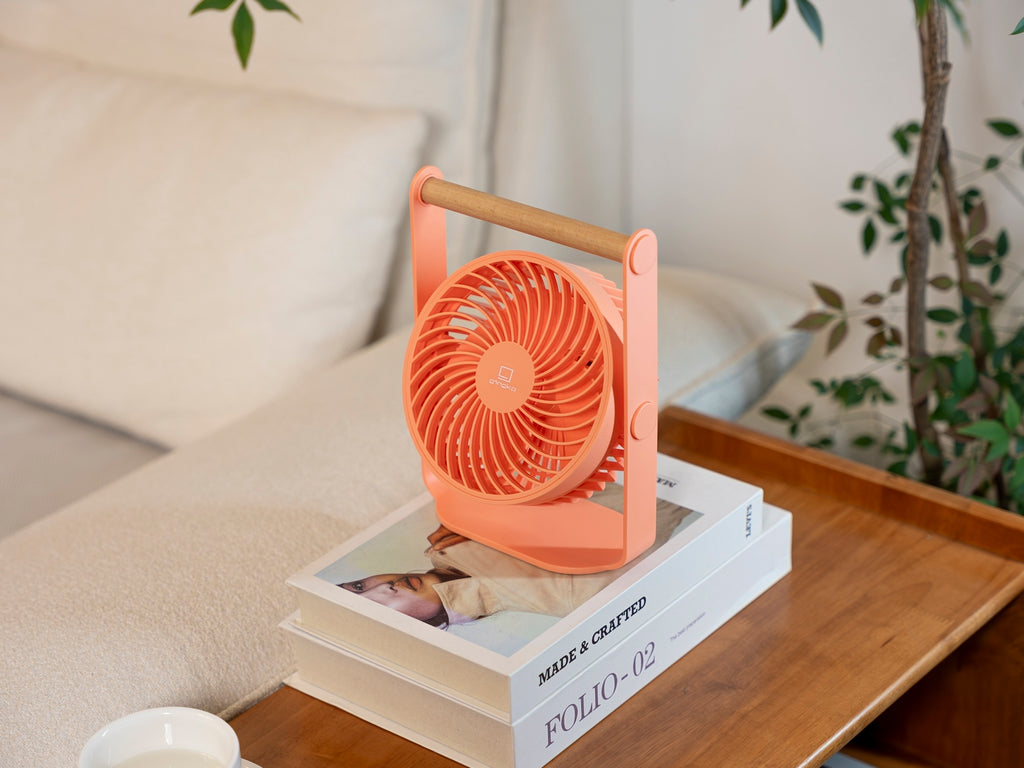 Gingko Design – Tragbarer Ventilator Lyra - Pfirsich, ein kleiner oranger Ventilator auf Büchern auf einem Holztisch stehend