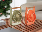 Gingko Design – Tragbarer Ventilator Lyra - Pfirsich, zwei tragbare Ventilatoren, einer grün und einer orange, auf einem Holztisch stehend