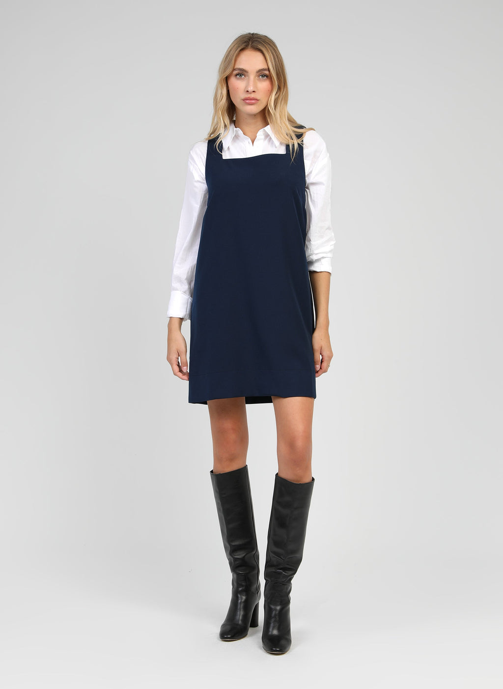 An'ge – Robe Courte Maleo Marine, mannequin blonde portant une robe chasuble bleu marine sur chemise blanche