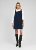 An'ge – Robe Courte Maleo Marine, mannequin blonde portant une robe chasuble bleu marine sur chemise blanche