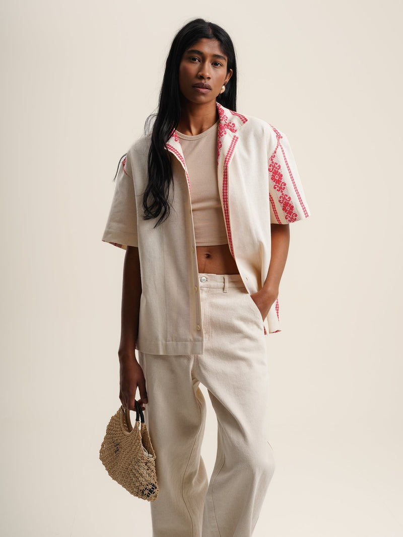 A model wearing a white shirt and pants with pink embroidery. The shirt is a red folklore short sleeve shirt.