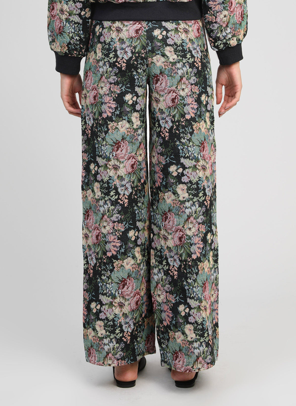 An'ge – Pantalon Passyna Noir, pantalon large noir à motif floral rose et vert, porté de dos