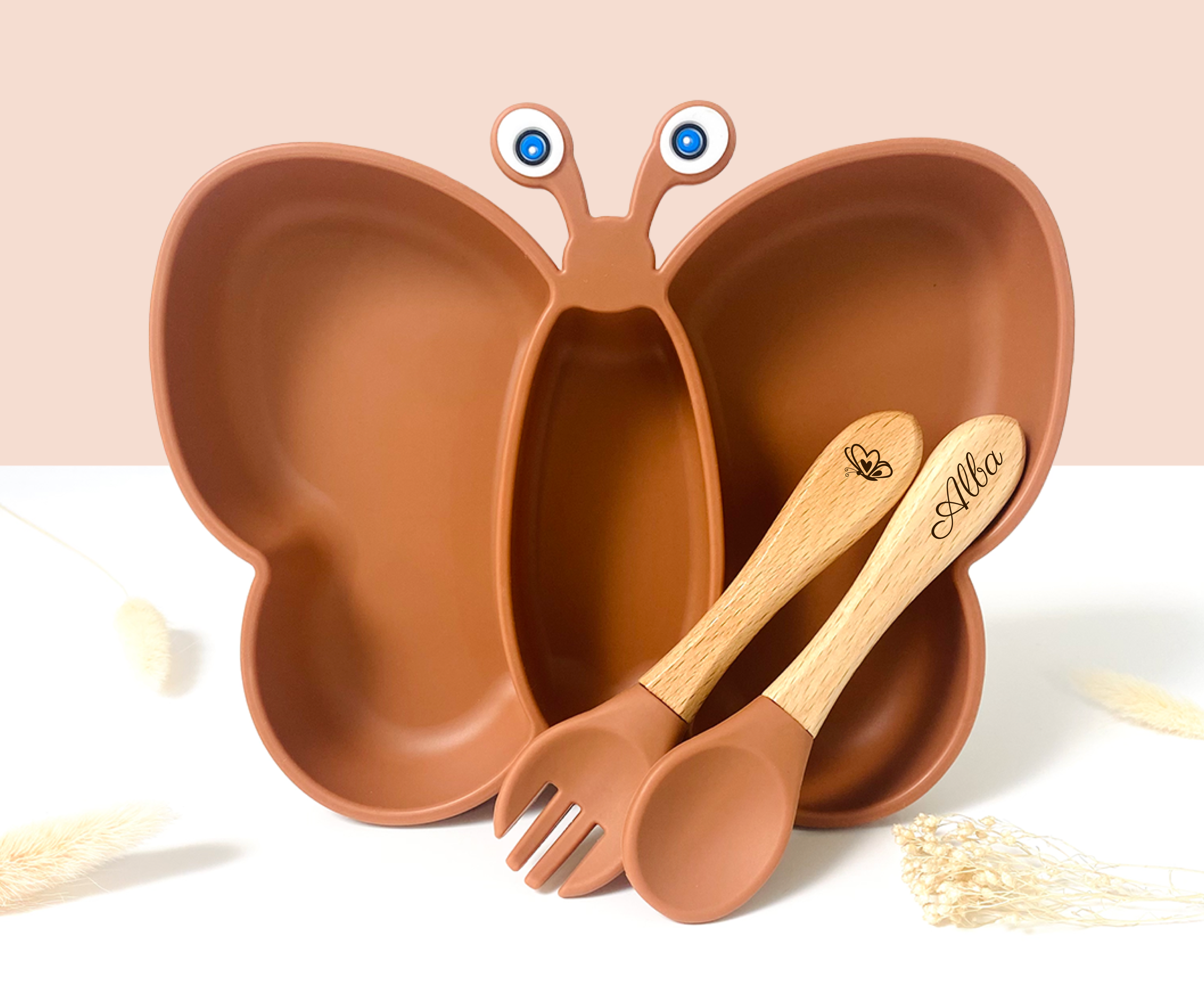 Assiette enfant en forme de papillon couleur terracotta avec antennes et yeux, accompagnée de couverts en bois gravés “Alba”, vue en plan rapproché.