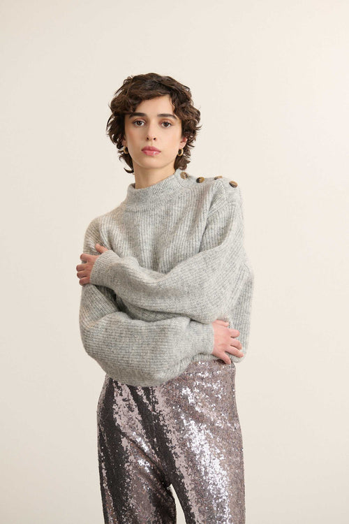 Mannequin femme de face portant un pull ample gris, col rond et ouverture boutonnée sur l’épaule avec boutons effet corne.