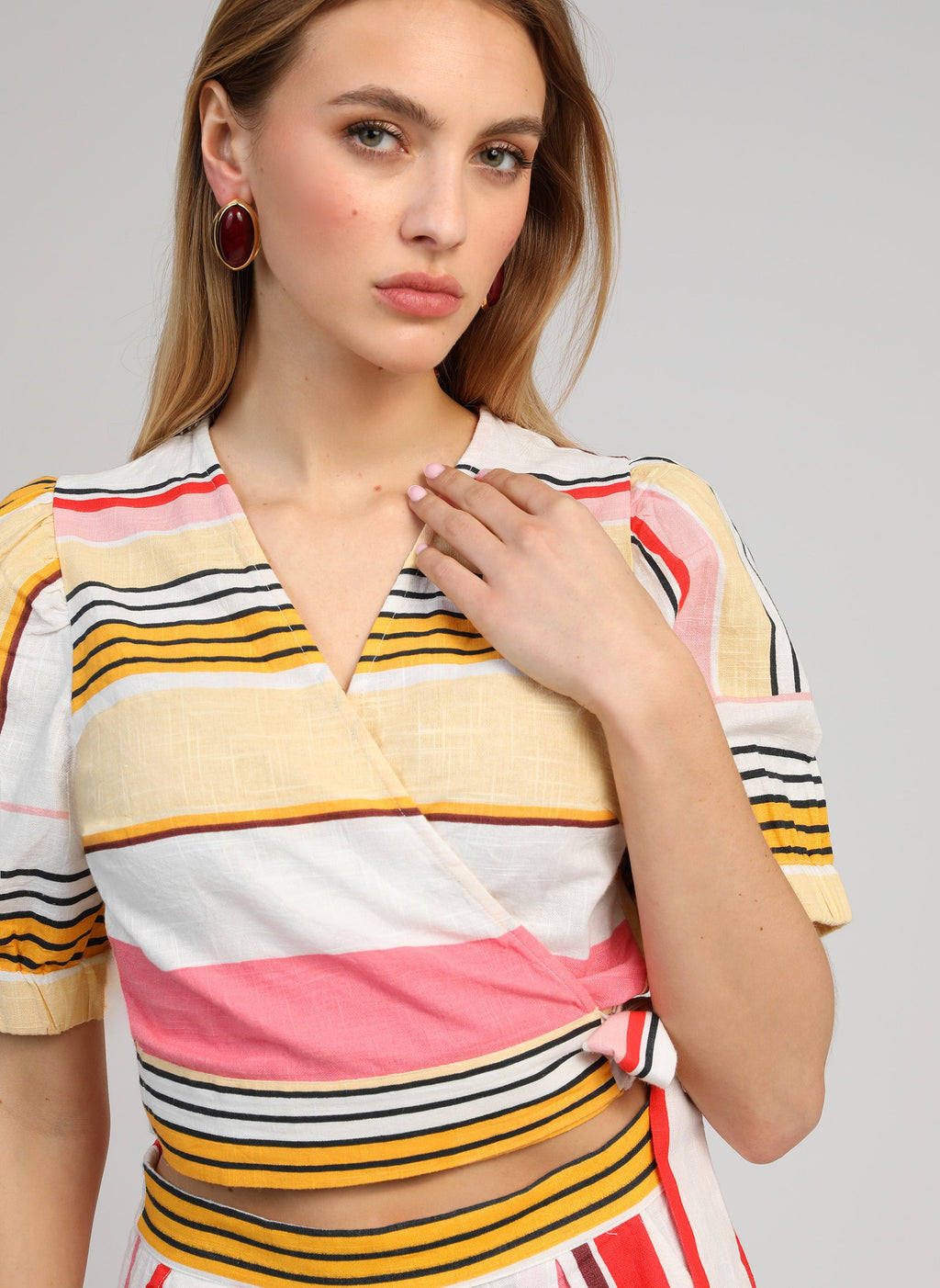 An'ge – Cache-Coeur Soriana Belinda Multico, model in een meerkleurig gestreepte crop top met rode oorbellen
