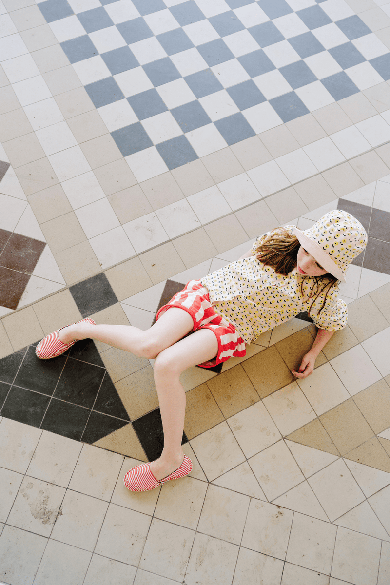 Fille portant le BOB à imprimé fleurs jaunes, la chemise SASHA à manches longues imprimé fleurs jaunes et le short KIM à rayures rouges, en coton bio style vintage mixte et unisexe