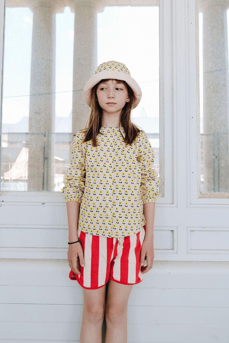 Fille portant le BOB à imprimé fleurs jaunes, la chemise SASHA à manches longues imprimé fleurs jaunes et le short KIM à rayures rouges, en coton bio style vintage mixte et unisexe