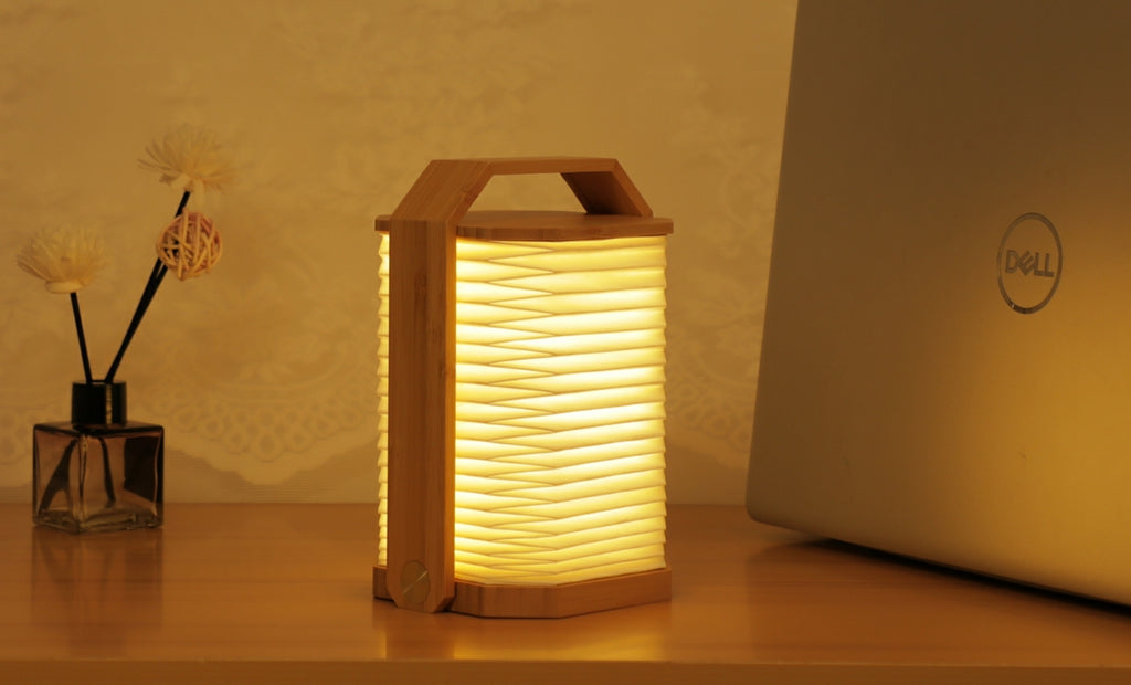 Gingko Design – Lampa Smart Origami - Naturalny Bambus, składana lampa z jasnego drewna, włączona, stojąca na drewnianym stole