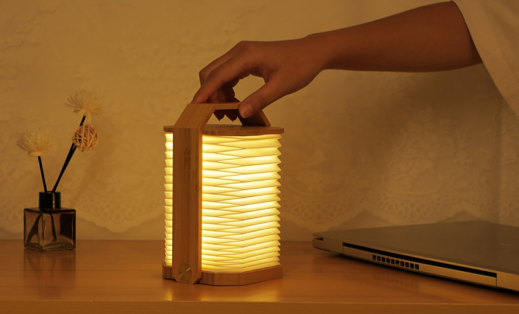 Gingko Design – Lampa Smart Origami - Naturalny Bambus, dłoń chwytająca włączoną lampę z drewna i plisowanego papieru