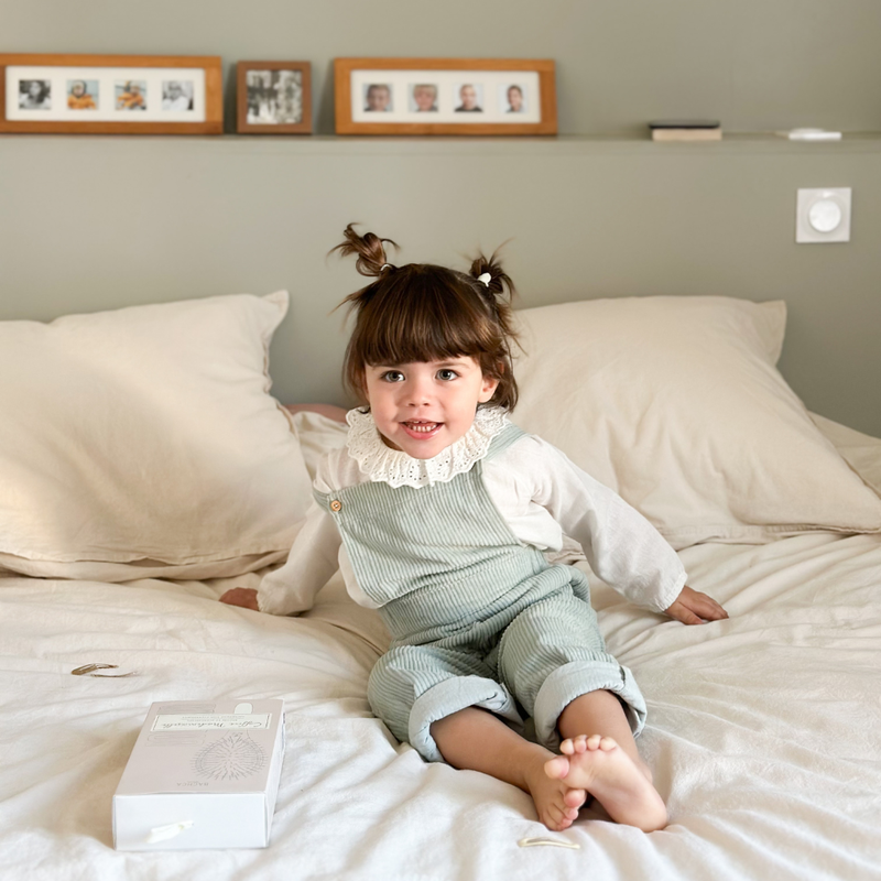 Petite fille sur un lit. Elle sourit à la caméra, on voit à côté d'elle un coffret d'accessoires cheveux de la marque Bachca.