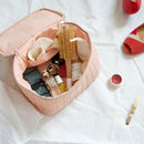 Trousse vanity Bachca en tissu rose poudré ouverte, contenant brosse ronde, pinceaux maquillage, barrettes, soins et accessoires beauté, posée sur un drap blanc.