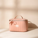 Vanity rectangulaire Bachca matelassé en coton rose poudré avec fermeture éclair dorée et petite anse, posé sur un fond beige lumineux.