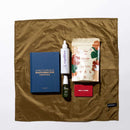 Ensemble des produits qui composent le coffret cadeau Noël Plaisir de Gapianne. Il y a un jeu de cartes, un livre éducatif, une infusion, une huile de massage pour le corps et une huile intime. Ils sont positionnés sur le furoshiki kaki Gapianne.