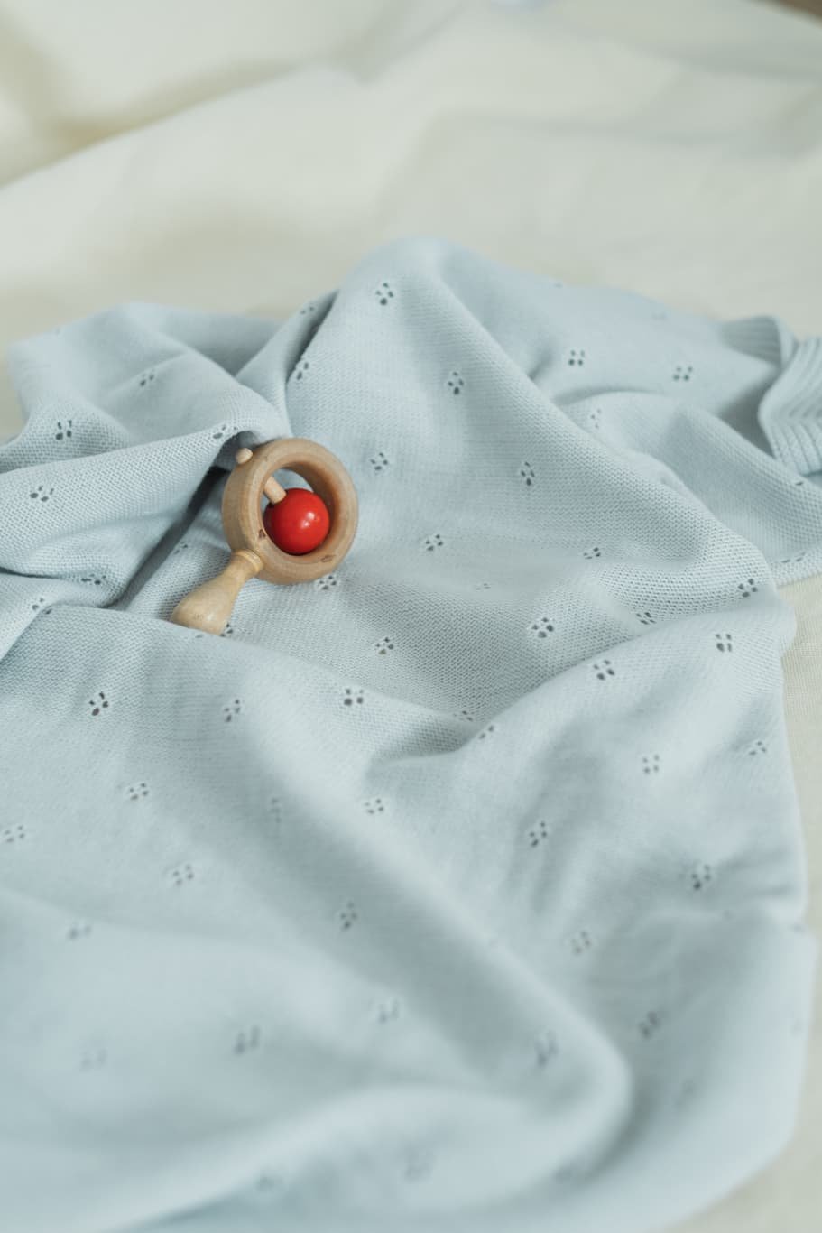 La Manufacture De Layette – Couverture Tricotée Ajourée - Vert D'Eau, un hochet en bois et rouge posé sur un tissu bleu clair