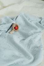 La Manufacture De Layette – Couverture Tricotée Ajourée - Vert D'Eau, un hochet en bois et rouge posé sur un tissu bleu clair