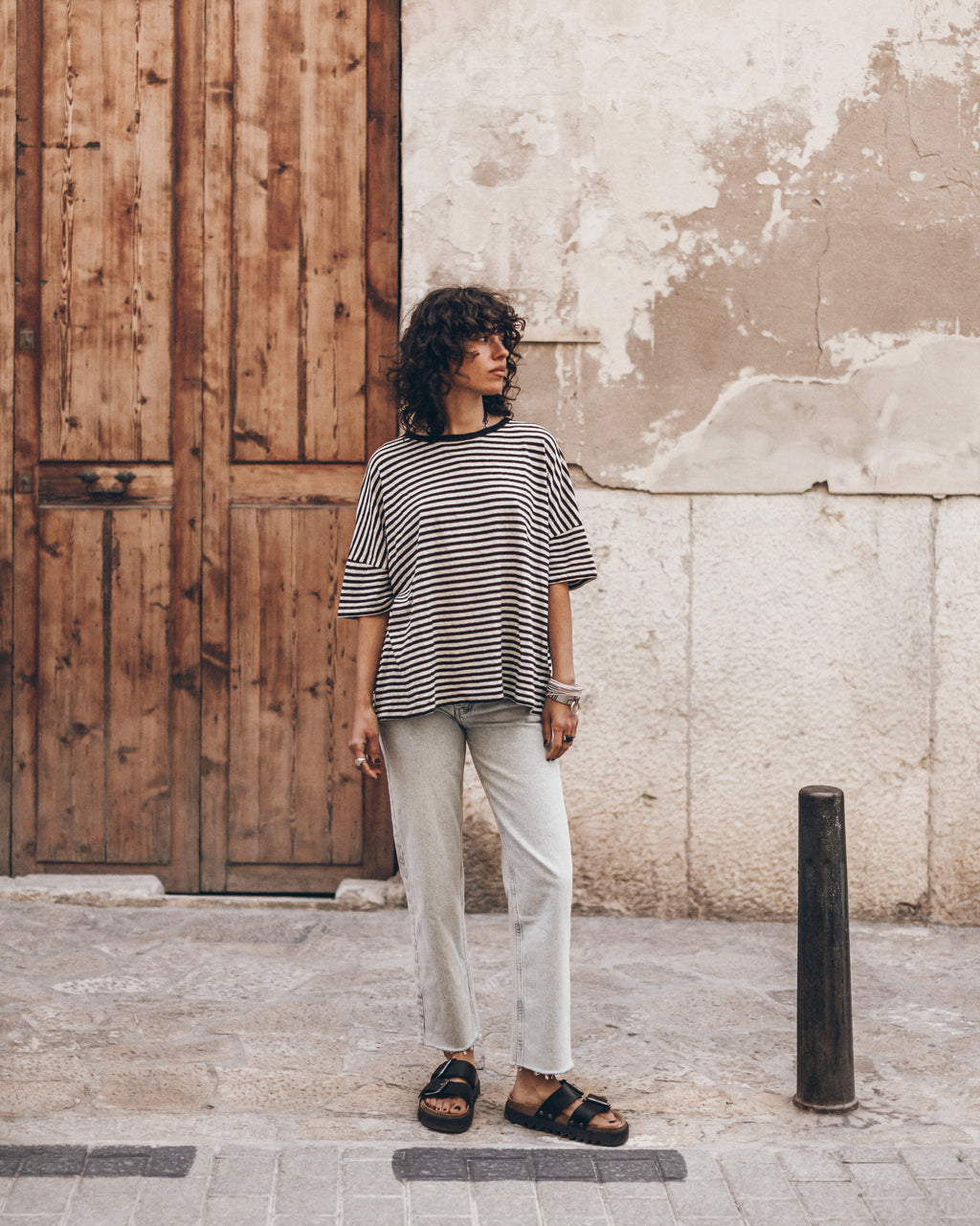 Woman standing on a street wearing a striped linen t-shirt, light jeans, and black leather sandals.