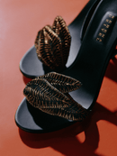 Close-up of elegant black high-heeled sandals with intricate leaf details on a vibrant orange background.