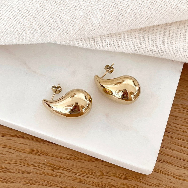Instants Plaisirs – Boucles D'Oreilles Gaia - Acier, boucles d'oreilles dorées en forme de goutte sur fond blanc