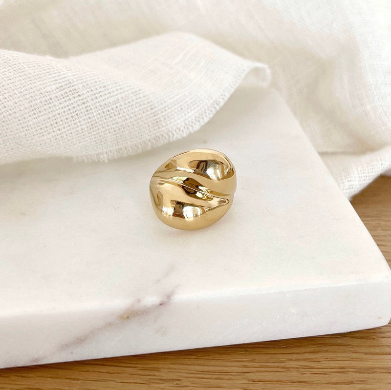 Instants Plaisirs – Bague Dody - Acier, bague dorée à la forme ondulée, posée sur un marbre blanc